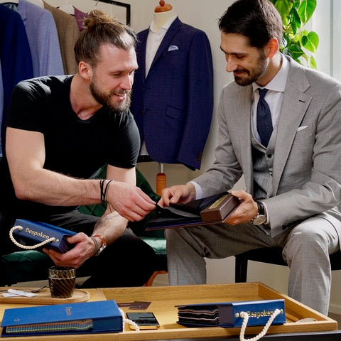 Two men in a suit store examining blue and brown wool fabrics, with branded blue bags on a wooden tray in the foreground. One man wears a light gray suit and the other wears a black t-shirt.